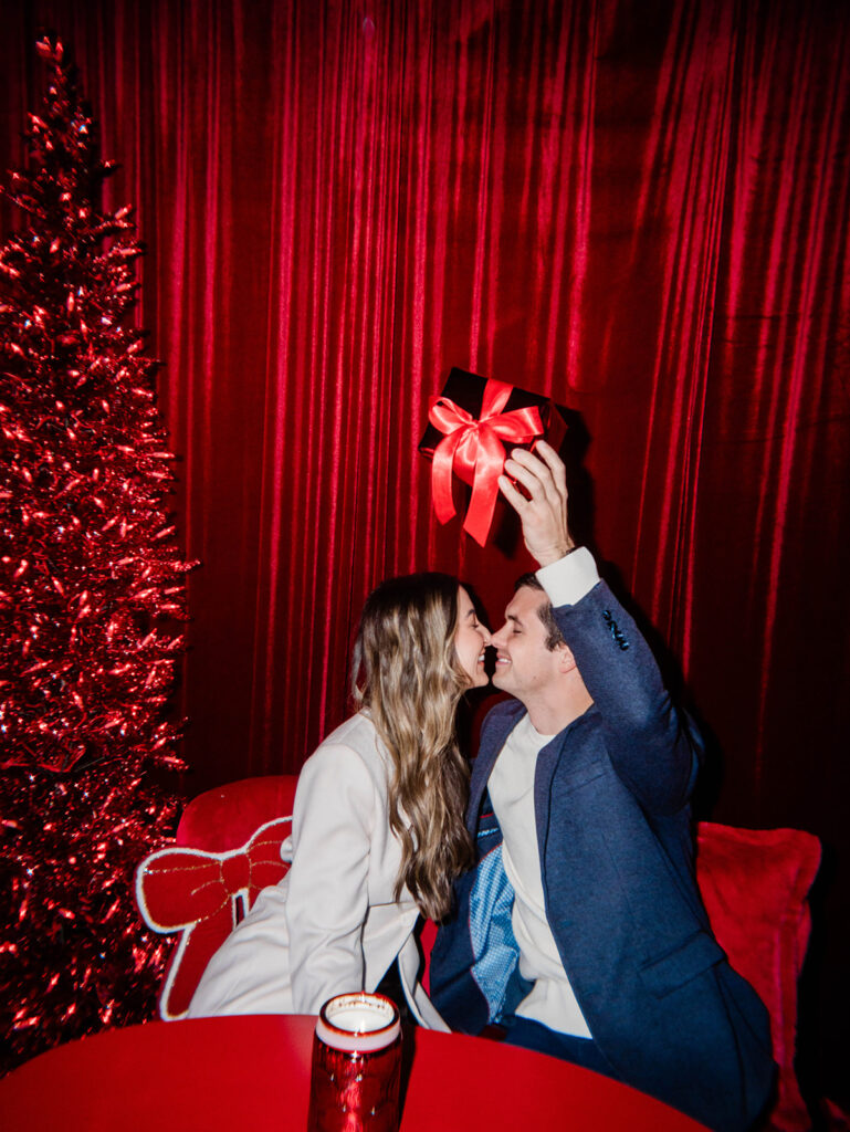 Couple celebrating at the Santa Baby pop-up bar, sitting beside a red tinsel tree with wrapped gifts, designed by Columbus event designers Aisle & Co.