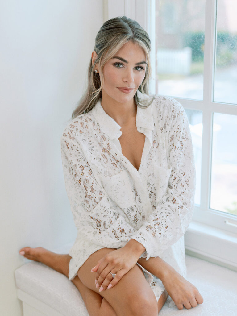 Bride in lace robe sitting by window in soft natural light.